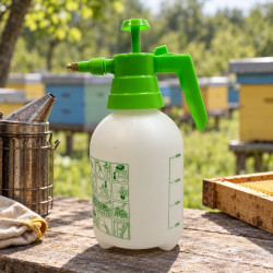 Sprayer on a wooden table near a smoker and frame, with an apiary in the background